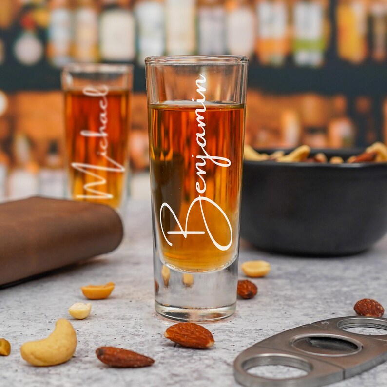 Op de afbeelding: Twee shotglaasjes gevuld met amberkleurige vloeistof, &eacute;&eacute;n met de tekst "Michael" en de andere "Benjamin" in witte letters. De glazen staan op een lichtgrijze ondergrond met verspreide noten, een zwarte kom met snacks en een metalen sigarenknipper.