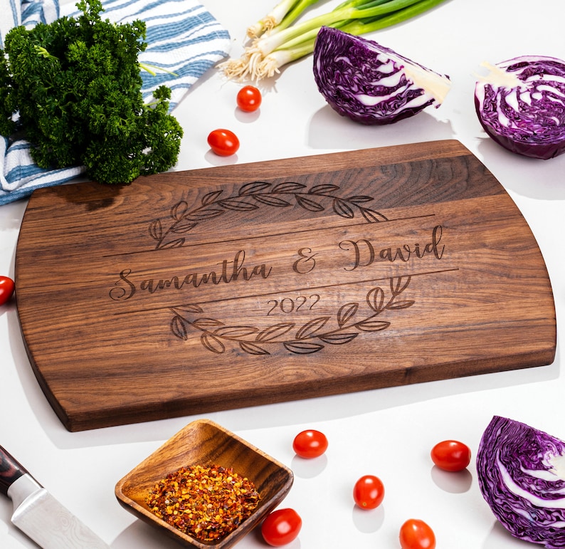 Puede incluir: Una tabla de cortar de madera de nogal personalizada con una corona grabada decorativa y los nombres "Samantha & David" y el a&ntilde;o "2022".