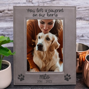 May include: A gray leather photo frame with a paw print design. The frame has a picture of a woman and a golden retriever dog. The text "You left a pawprint on our hearts" is engraved on the frame. The dog's name "Milo" and the dates "2014 - 2023" are also engraved on the frame.
