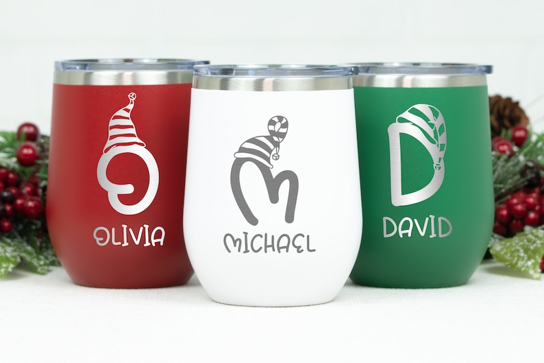 May include: Three stainless steel wine tumblers with personalized names and a Christmas-themed design. The tumblers are red, white, and green, and each features a different letter with a Santa hat. The names on the tumblers are Olivia, Michael, and David.