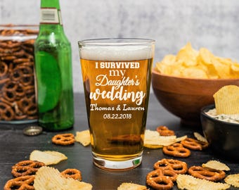 May include: A pint glass filled with beer, featuring the text "I Survived My Daughter's Wedding" with the names Thomas & Lauren and the date 08.22.2018. The glass is surrounded by snacks, including pretzels, potato chips, and a green beer bottle.