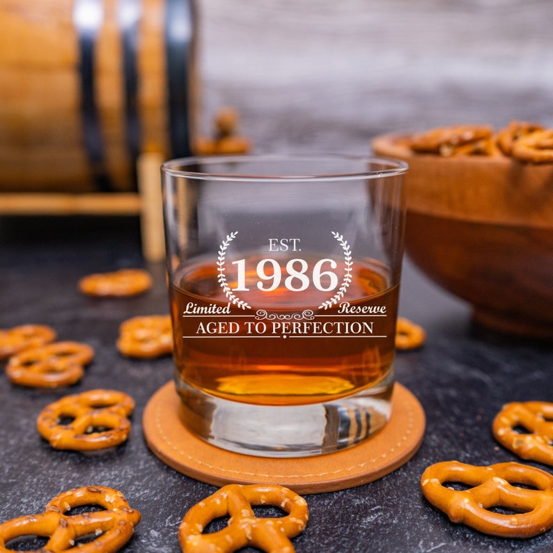 May include: A clear glass with a brown liquid inside. The glass has a white design that says "EST. 1986 Limited Reserve Aged to Perfection".