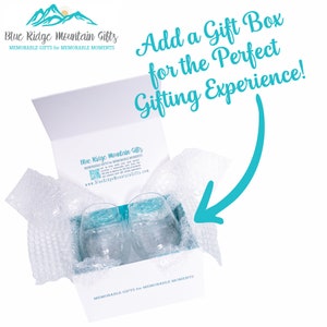 May include: A white gift box with a blue and white logo that says "Blue Ridge Mountain Gifts" and "MEMORABLE GIFTS for MEMORABLE MOMENTS". The box is open and contains two clear glass wine glasses wrapped in bubble wrap. The text "Add a Gift Box for the Perfect Gifting Experience!" is written in blue cursive on a white background.