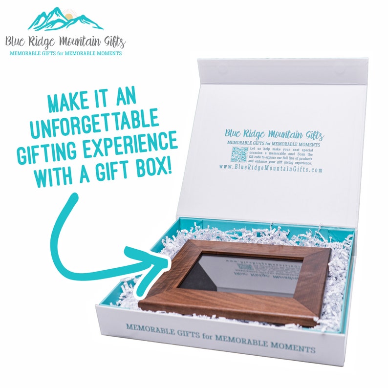 K&ouml;nnte beinhalten: Ein Holzbildrahmen in einer wei&szlig;en Geschenkbox mit blau-wei&szlig;em Seidenpapier. Die Box ist ge&ouml;ffnet und der Rahmen ist sichtbar. Der Text auf der Box lautet "Blue Ridge Mountain Gifts, Denkw&uuml;rdige Geschenke f&uuml;r denkw&uuml;rdige Momente".