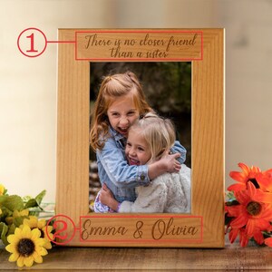 May include: A wooden picture frame with a photo of two young girls hugging. The frame has an inscription that reads "There is no closer friend than a sister." and the names "Emma & Olivia" are engraved below the photo.