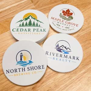 May include: Four round white coasters featuring distinct logos. One shows Cedar Peak Outfitters with a mountain, one Maple Grove Market with a maple leaf, one North Shore Brewing Co. with a lighthouse, and one Rivermark Realty with a house.