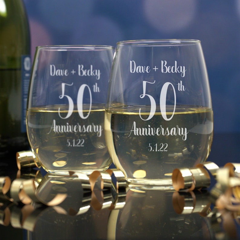 May include: Two clear glass wine glasses with the text "Dave + Becky 50th Anniversary 5.1.22" etched on them.