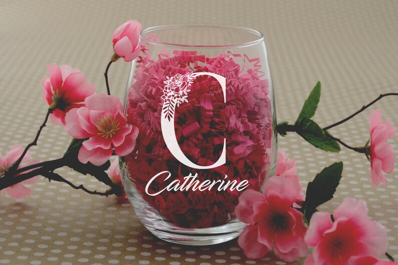 Puede incluir: Una copa de vino de vidrio transparente con un dise&ntilde;o floral blanco y el nombre "Catherine" grabado en el lateral. La copa est&aacute; llena de papel de seda rosa.