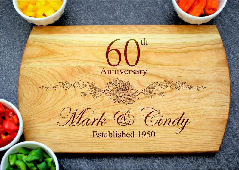K&ouml;nnte beinhalten: Ein Schneidebrett aus Holz mit einer 60. Jahrestag-Inschrift, einem floralen Design und den Namen "Mark & Cindy" mit dem Jahr "1950" eingraviert.