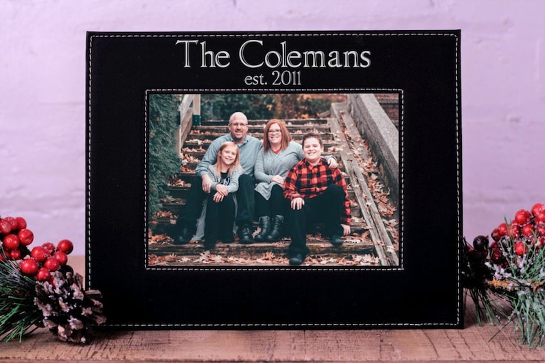 K&ouml;nnte beinhalten: Schwarzer Leder-Bilderrahmen mit einem gen&auml;hten Rand. Der Rahmen hat ein Foto von einer vierk&ouml;pfigen Familie, die auf Stufen sitzt. Der Text "The Colemans est. 2011" ist auf dem Rahmen gedruckt.