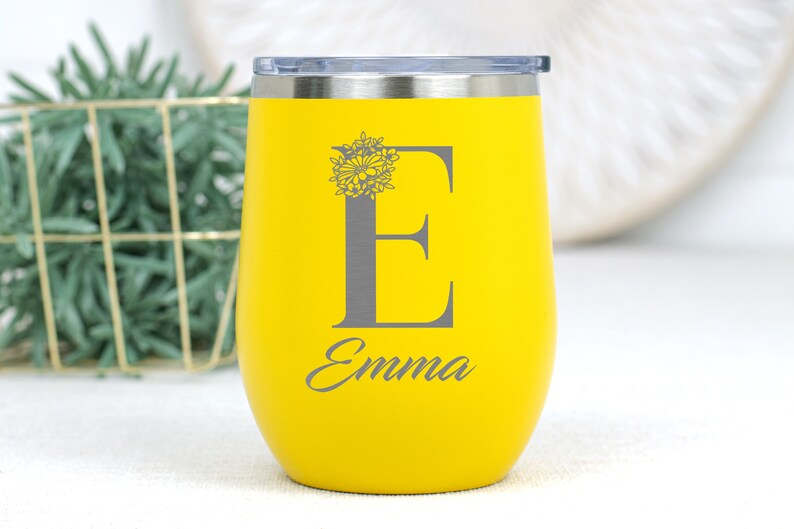K&ouml;nnte beinhalten: Gelber Edelstahl-Weinglas mit einem Blumendesign und dem Buchstaben "E" auf der Vorderseite graviert. Der Name "Emma" ist unter dem Buchstaben graviert.