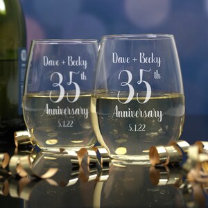 May include: Two clear glass wine glasses with the text "Dave + Becky 35th Anniversary 5.1.22" etched on the front. The glasses are filled with white wine.