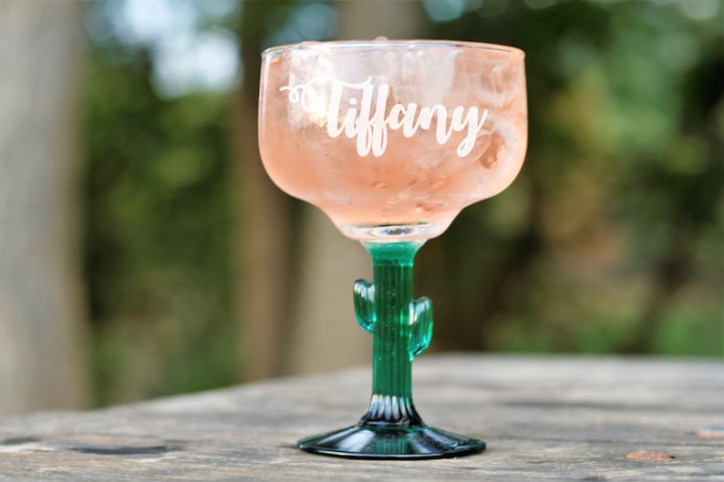 Op de afbeelding: Een groen glazen margarita-glas met een cactussteel en -voet. Het glas is gevuld met een roze drankje en ijs. Het glas is gepersonaliseerd met de naam "Tiffany".