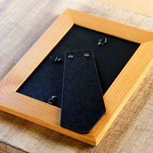 May include: A wooden picture frame with a black backing and a black felt easel stand. The frame has a natural wood finish.