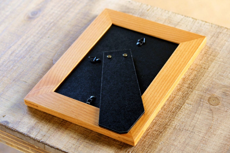 May include: A wooden picture frame with a black backing and a black felt easel stand. The frame has a natural wood finish.