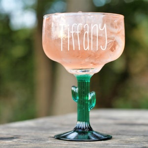 May include: A clear glass margarita glass with a green cactus stem and base. The glass is filled with a pink drink and ice. The word "Tiffany" is written on the side of the glass.