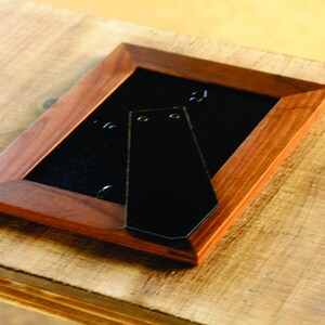 Pu&ograve; includere: Una cornice per foto in legno marrone scuro con un retro nero e un gancio di metallo per appendere. La cornice &egrave; su una superficie in legno marrone chiaro.