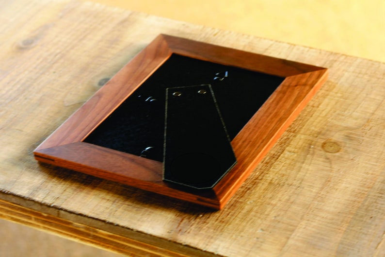 Pu&ograve; includere: Una cornice per foto in legno marrone scuro con un retro nero e un gancio di metallo per appendere. La cornice &egrave; su una superficie in legno marrone chiaro.
