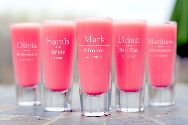May include: Five clear glass shot glasses with pink liquid. Each glass is engraved with a name and title, such as "Olivia Bridesmaid" and "Mark Groom".