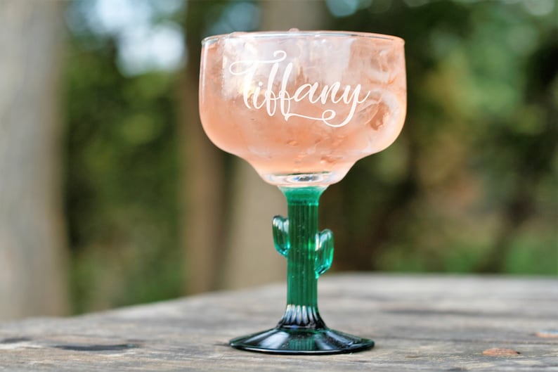 May include: A clear glass margarita glass with a green cactus stem and base. The glass is filled with a pink drink and ice. The word "Tiffany" is etched on the side of the glass.