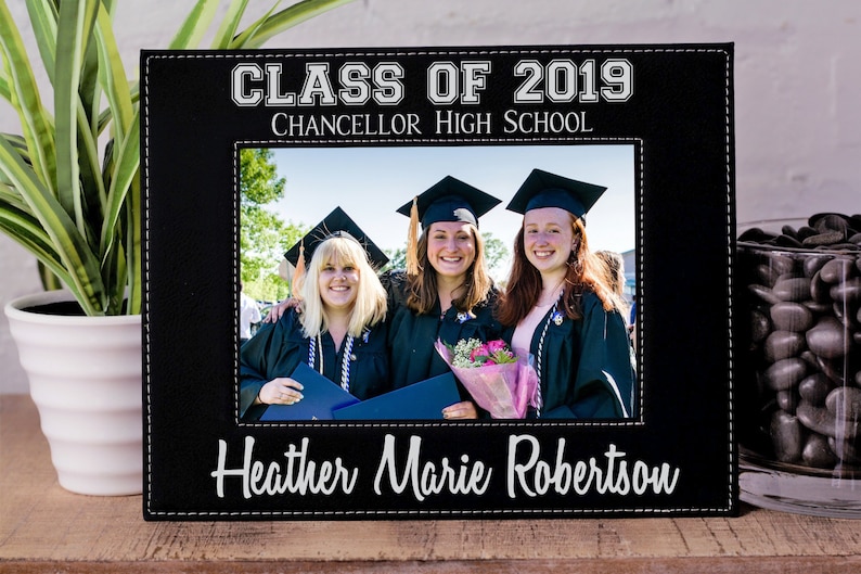 K&ouml;nnte beinhalten: Schwarzer Lederbilderrahmen mit wei&szlig;en N&auml;hten. Der Rahmen enth&auml;lt ein Foto von drei Personen in Abschlussroben. Der Text auf dem Rahmen lautet "Klasse von 2019 Chancellor High School Heather Marie Robertson".