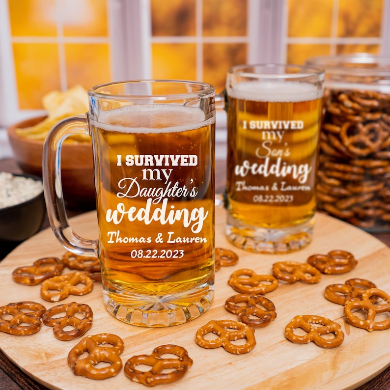 May include: Two clear glass beer mugs with handles. The mugs are filled with a golden-colored liquid. The mugs are decorated with white text that reads "I survived my daughter's wedding" and "I survived my son's wedding". The text also includes the names "Thomas & Lauren" and the date "08.22.2023".
