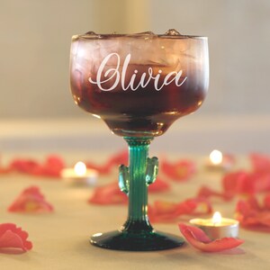 May include: A clear glass margarita glass with a green cactus stem and base. The glass is filled with a red drink and ice. The glass is personalized with the name "Olivia" in white lettering.