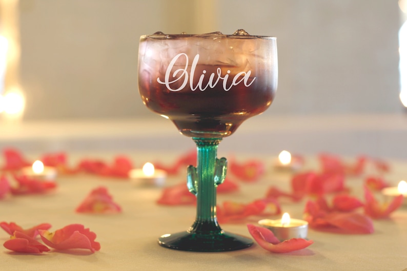 May include: A clear glass margarita glass with a green cactus stem and base. The glass is filled with a red drink and ice. The glass is personalized with the name "Olivia" in white lettering.