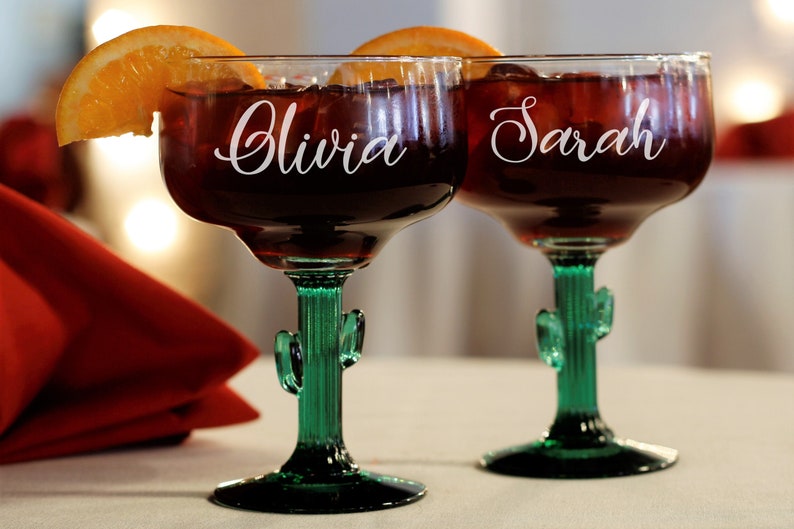 May include: Two green cactus-shaped margarita glasses with the names "Olivia" and "Sarah" etched on them. The glasses are filled with a red beverage and garnished with orange slices.