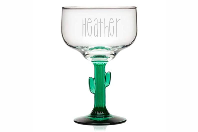 May include: A clear glass margarita glass with a green cactus stem and base. The glass is engraved with the name "Heather".