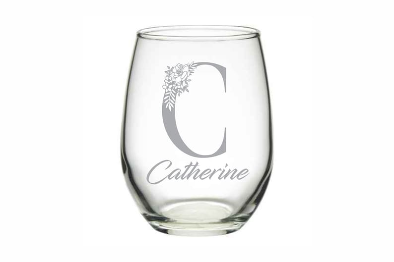 Puede incluir: Una copa de vino sin tallo de vidrio transparente con un dise&ntilde;o floral gris y la letra "C" y el nombre "Catherine" grabados en la parte delantera.