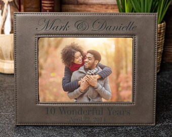 Può includere: Una cornice per foto in pelle marrone con un bordo cucito. La cornice contiene una foto di una coppia che sorride e si abbraccia. La cornice è incisa con il testo "Mark & Danielle" e "10 Wonderful Years April 22, 2014".