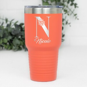 K&ouml;nnte beinhalten: Ein korallenfarbener Edelstahlbecher mit einem wei&szlig;en Blumenmuster und dem Buchstaben "N" sowie dem Namen "Nicole" auf der Seite graviert.