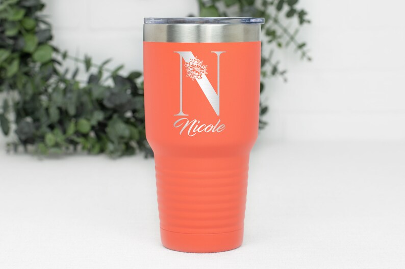 K&ouml;nnte beinhalten: Ein korallenfarbener Edelstahlbecher mit einem wei&szlig;en Blumenmuster und dem Buchstaben "N" sowie dem Namen "Nicole" auf der Seite graviert.