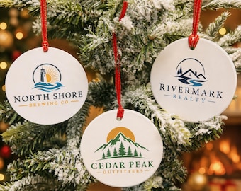 May include: Three round white ceramic ornaments with red ribbon hangers. The left ornament features a lighthouse logo and the text NORTH SHORE BREWING CO. The right ornament shows a house and river logo with RIVERMARK REALTY. The bottom ornament displays a mountain and pine tree logo with CEDAR PEAK OUTFITTERS.
