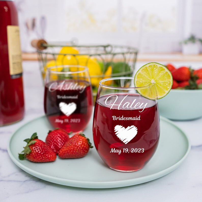 May include: Two wine glasses with red wine. The glasses are engraved with the names "Ashley" and "Haley" and the text "Bridesmaid" and "May 19, 2023".
