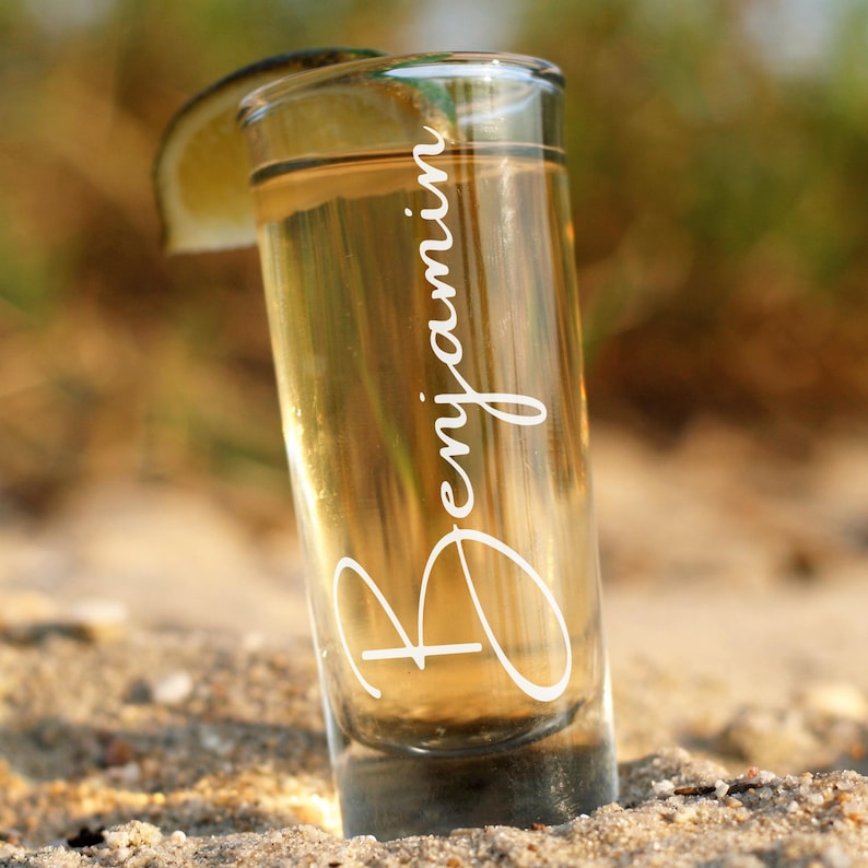 Op de afbeelding: Een helder shotglaasje gevuld met een gouden vloeistof, mogelijk tequila, en een schijfje limoen. De naam "Benjamin" staat in elegant wit schrift op het glas. Het glas staat in het zand.