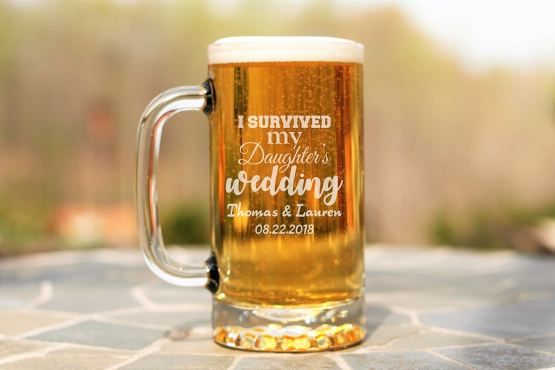 May include: A clear glass beer mug with a handle. The mug is filled with a golden-colored beer and has the text "I survived my daughter's wedding Thomas & Lauren 08.22.2018" etched on the side.