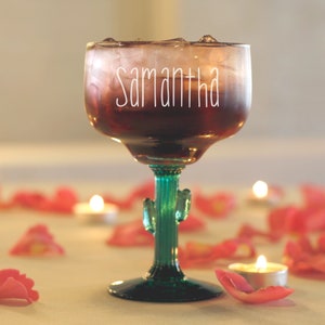 May include: A clear glass margarita glass with a cactus-shaped stem, filled with a dark red liquid and ice. The glass has the name "Samantha" written on it in white. The glass is surrounded by pink rose petals and small white candles.
