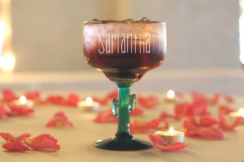 May include: A clear glass margarita glass with a cactus-shaped stem, filled with a dark red liquid and ice. The glass has the name "Samantha" written on it in white. The glass is surrounded by pink rose petals and small white candles.