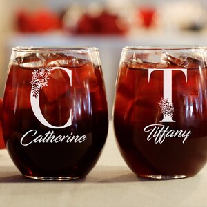 Puede incluir: Dos copas de vino transparentes con vino tinto. Las copas est&aacute;n personalizadas con los nombres "Catherine" y "Tiffany" y sus iniciales "C" y "T" con dise&ntilde;os florales.