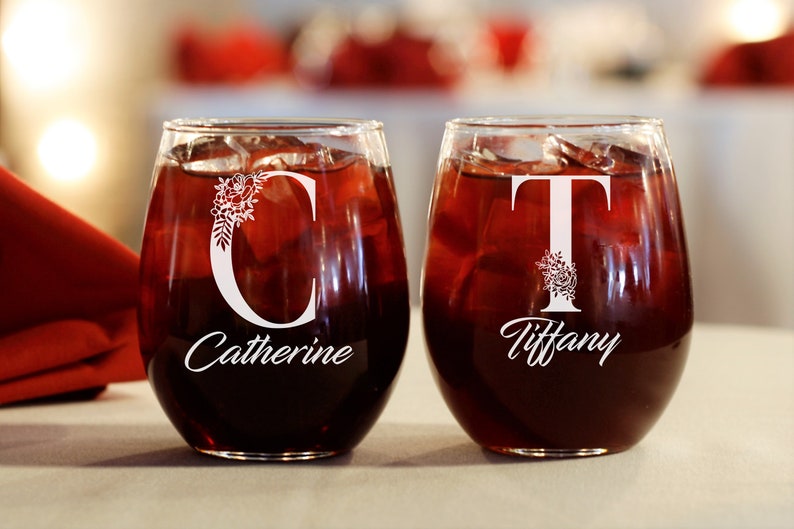 Puede incluir: Dos copas de vino transparentes con vino tinto. Las copas est&aacute;n personalizadas con los nombres "Catherine" y "Tiffany" y sus iniciales "C" y "T" con dise&ntilde;os florales.