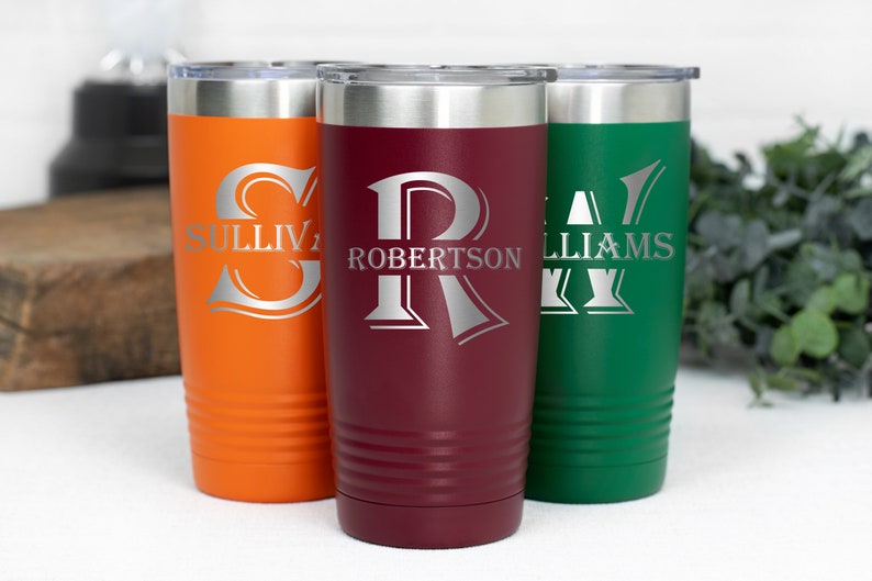 May include: Three personalized stainless steel tumblers in orange, burgundy, and green. Each tumbler features a silver monogram and last name: "Sullivan", "Robertson", and "Williams". The tumblers have a silver lid and a ridged design near the base.