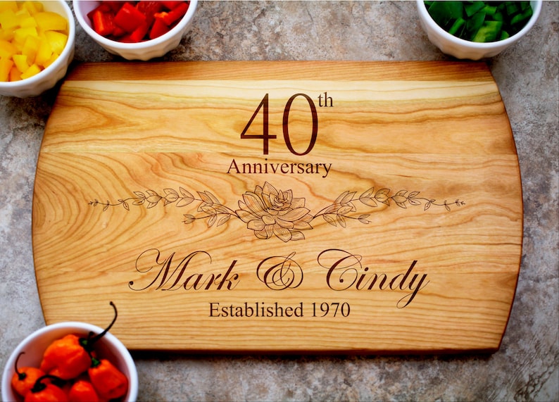 May include: A wooden cutting board engraved with "40th Anniversary", "Mark & Cindy", and "Established 1970". The board has a floral design and a natural wood grain.