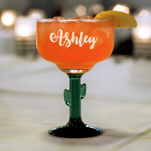 Op de afbeelding: Een groen cactusvormig margarita-glas met de naam "Ashley" gegraveerd aan de zijkant, gevuld met een roze drankje en een citroenwig als garnering.