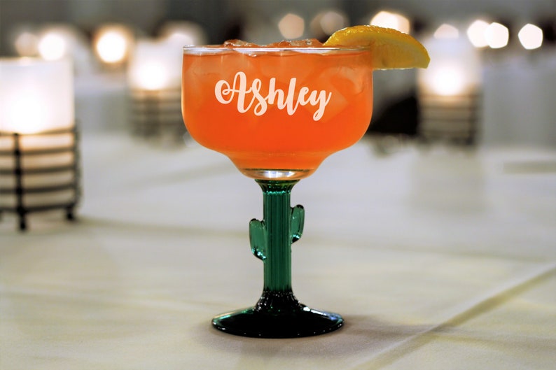 Op de afbeelding: Een groen cactusvormig margarita-glas met de naam "Ashley" gegraveerd aan de zijkant, gevuld met een roze drankje en een citroenwig als garnering.
