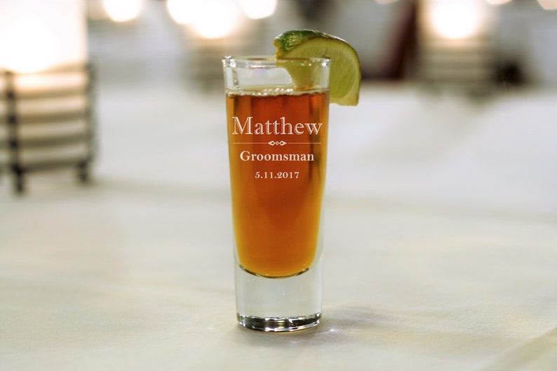 May include: A clear glass shot glass with a brown liquid inside. The glass is engraved with the text "Matthew Groomsman 5.11.2017". A lime wedge is on top of the glass.