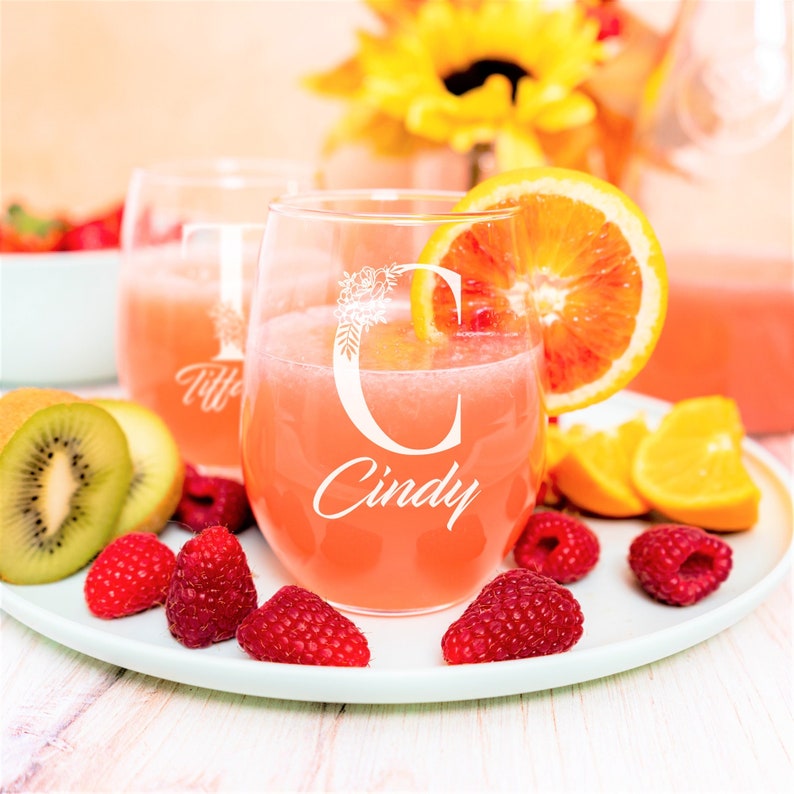 Puede incluir: Dos copas de vino sin tallo con nombres personalizados, "Tiff" y "Cindy", con dise&ntilde;os florales. Las copas est&aacute;n llenas de una bebida rosa y rodeadas de fruta, incluyendo frambuesas, kiwi y rodajas de naranja.