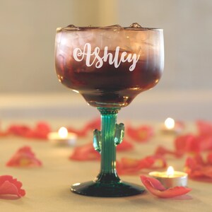 Op de afbeelding: Een helder glazen margarita-glas met een groene cactussteel en -voet. Het glas is gevuld met een rode vloeistof en ijs. Het glas is gepersonaliseerd met de naam "Ashley" in witte letters.
