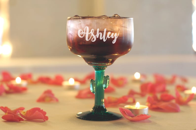 Op de afbeelding: Een helder glazen margarita-glas met een groene cactussteel en -voet. Het glas is gevuld met een rode vloeistof en ijs. Het glas is gepersonaliseerd met de naam "Ashley" in witte letters.